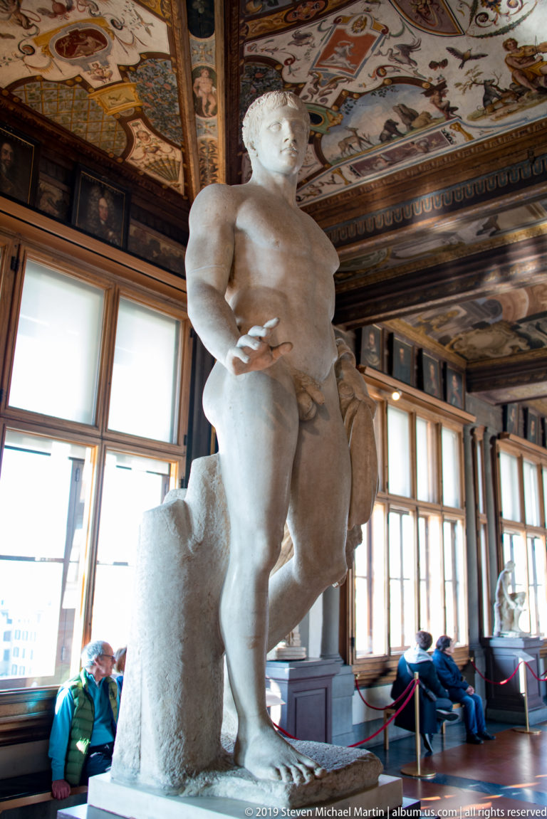 Nude marble male statue beside a tall rough support in a windowed Uffizi corridor with a painted ceiling.