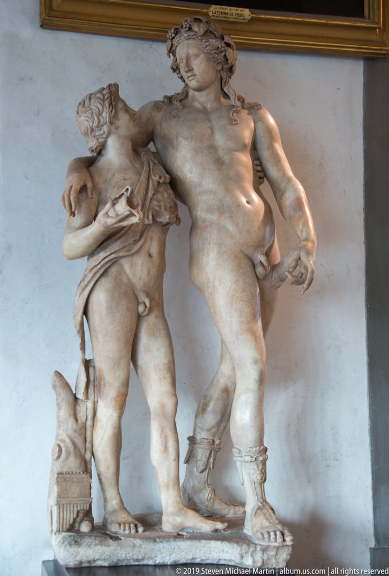 Two nude marble male figures stand together on one base, with drapery and carved support forms beside them.
