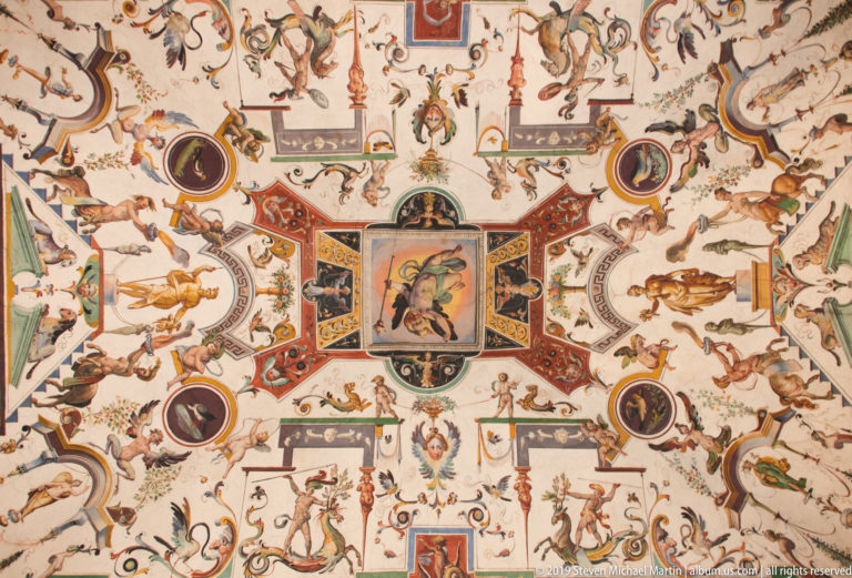 Symmetrical painted ceiling with grotesques, animals, medallions, small narrative panels, and red-green geometric borders.