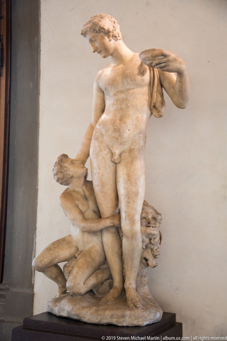 Nude marble female figure turns in contrapposto above a crouching sculptural support, with drapery over one shoulder.