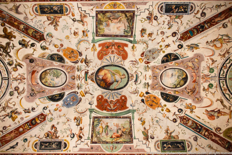 Ornate ceiling covered with painted grotesques, foliage, masks, medallions, and framed narrative scenes.