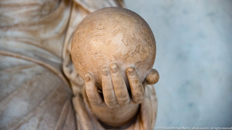Close view of a mottled stone globe with faint markings resting in the carved hand of a marble statue.