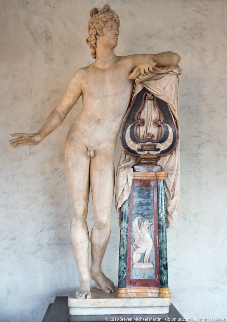 Nude marble female figure leans beside an ornate draped support with colored stone and a small inset figure.
