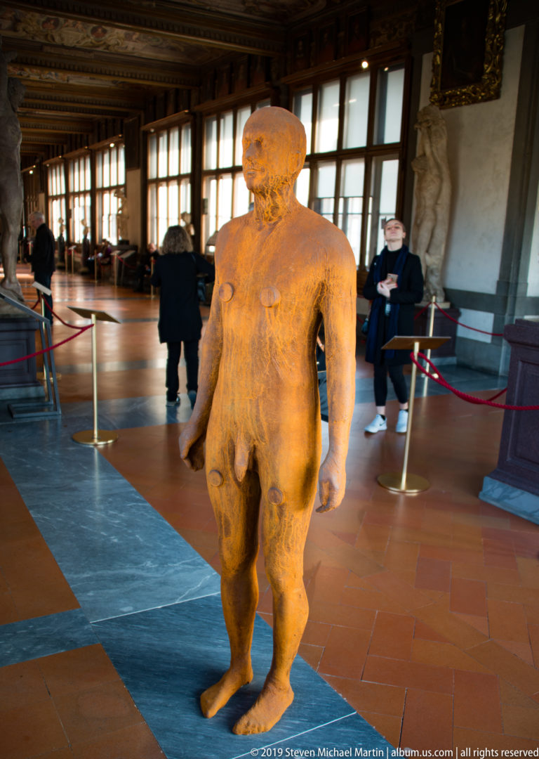 Standing nude marble male statue in a long Uffizi corridor with windows, visitors, and other sculptures.