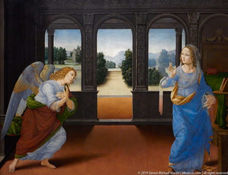 Kneeling angel faces the standing Virgin across a dark interior with three arches opening onto a landscape.