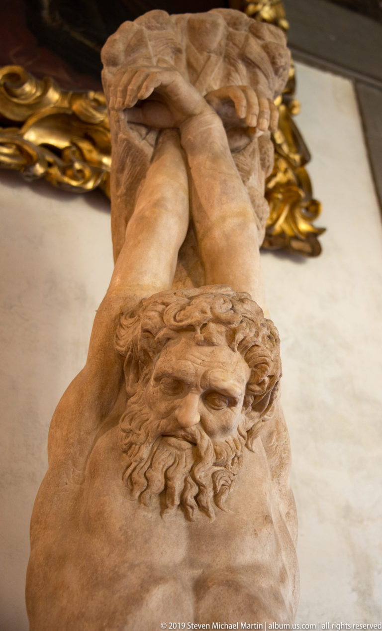 Upside-down close view of a bearded marble head belonging to a reclining nude sculptural figure.