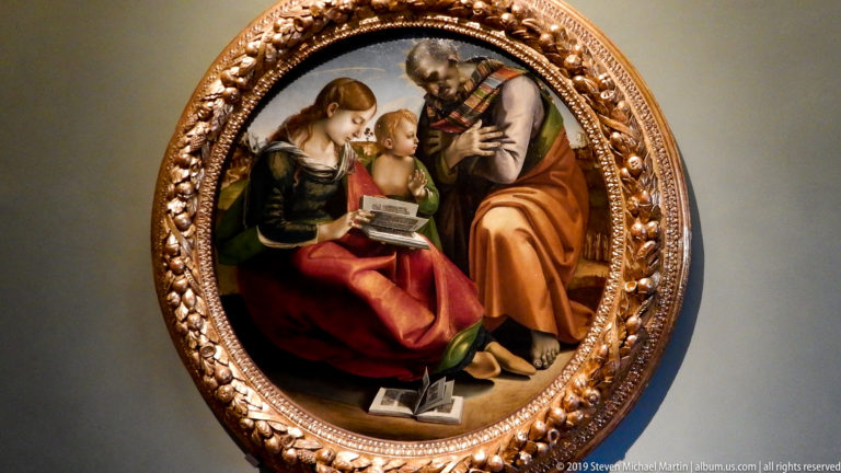 Circular painting of Mary, the Christ Child, and an older bearded man gathered around an open book.