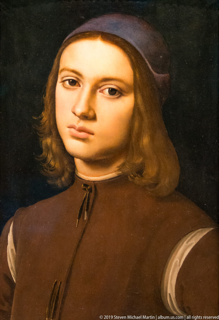 Young man with shoulder-length light brown hair wears a dark cap and brown doublet against a black background.