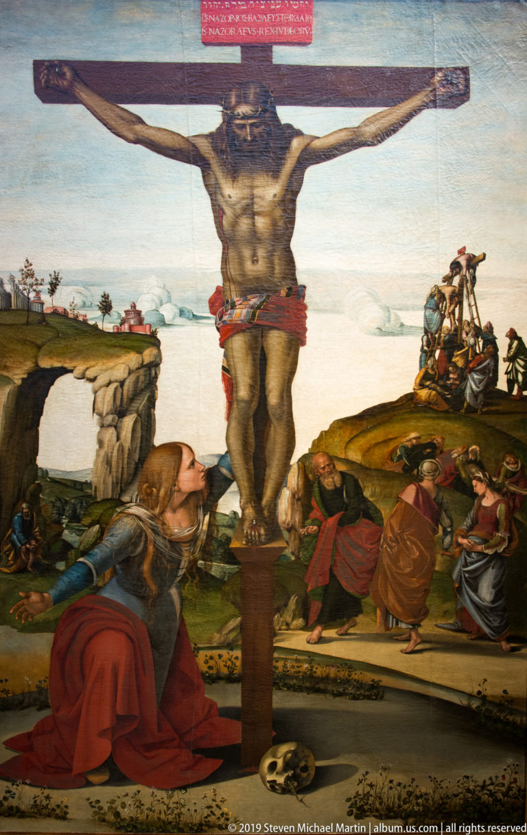 Christ crucified above kneeling mourners, with mounted figures, rocky terrain, and a distant settlement across the landscape.