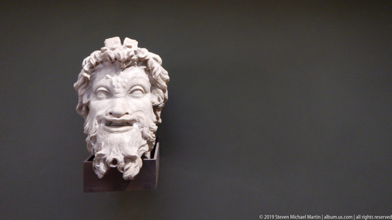 Small marble head with raised brows, open smiling mouth, short curls, and a full curling beard on a dark wall.