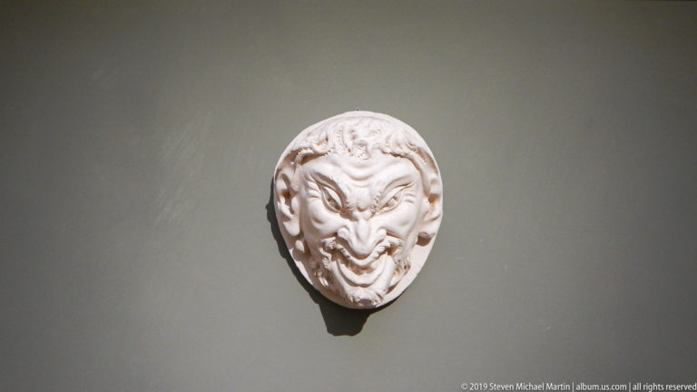 Small white carved mask with pointed ears, arched brows, curling facial forms, and a grimacing expression on a gray wall.