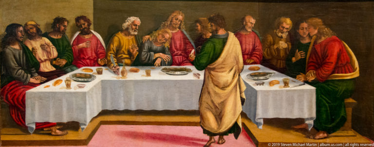 Christ and apostles sit around a long white table with bread and dishes, with one figure standing in the foreground.