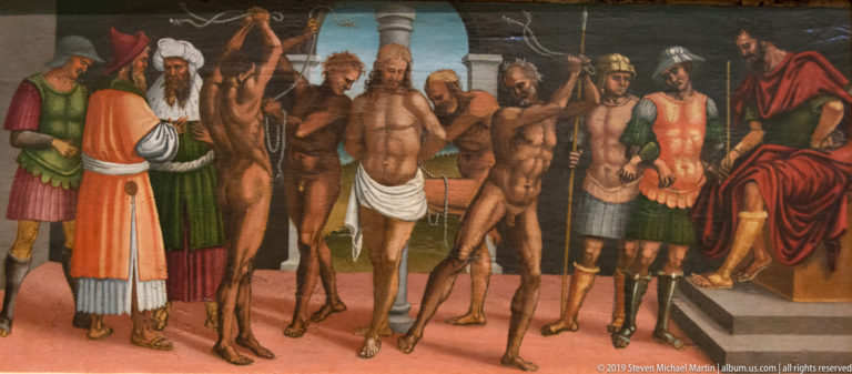 Nearly nude men stand among armed and richly dressed figures before an elevated man in a crowded narrative painting.