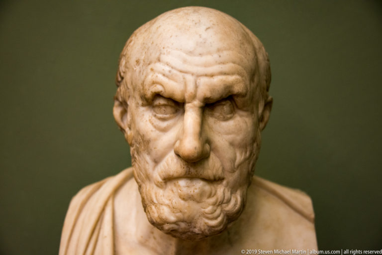 Frontal marble bust of a bald older man with knitted brow, sparse beard, and drapery over one shoulder.