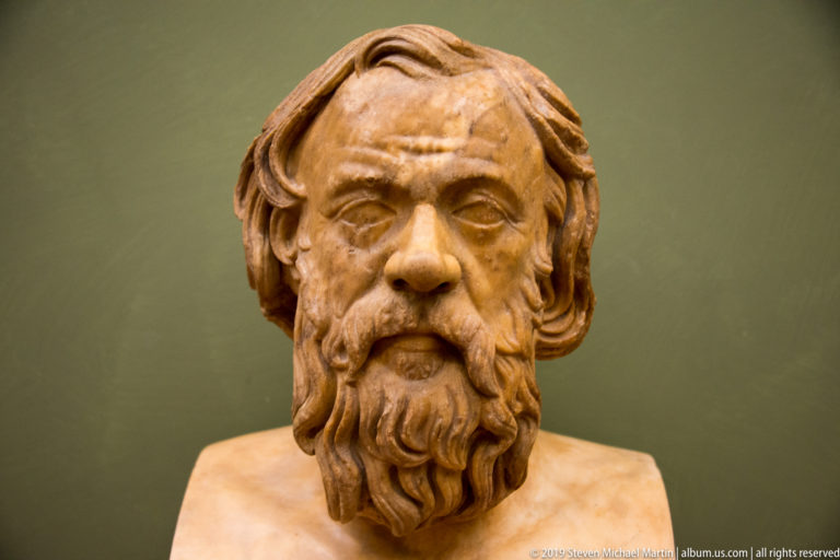 Frontal marble bust of a mature man with shoulder-length parted hair and a long deeply carved beard.