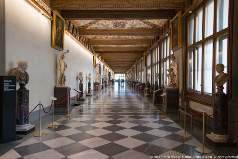 Long Uffizi corridor with checkered floor, classical statues, busts, tall windows, red ropes, and an ornate painted ceiling.