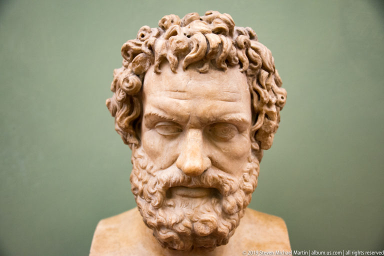 Frontal marble bust of a bearded man with dense tightly curled hair and deeply drilled beard.