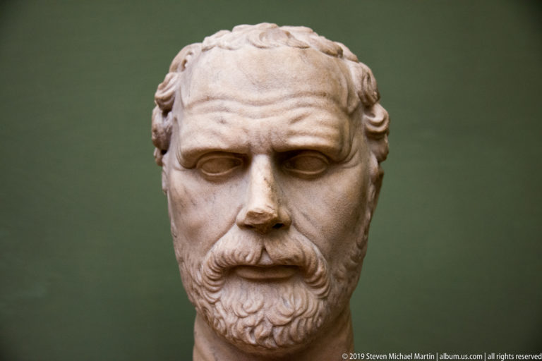 Frontal marble portrait of a short-haired man with a heavy brow, mustache, and compact beard.