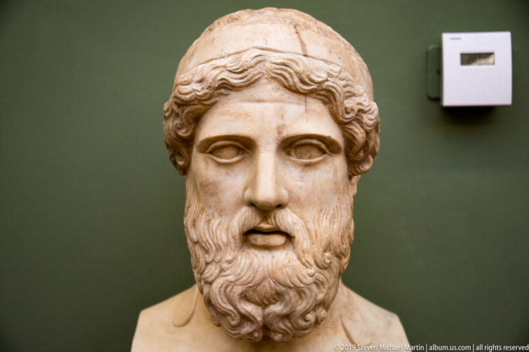 Frontal marble bust of a solemn man with center-parted wavy hair and a long curled beard.