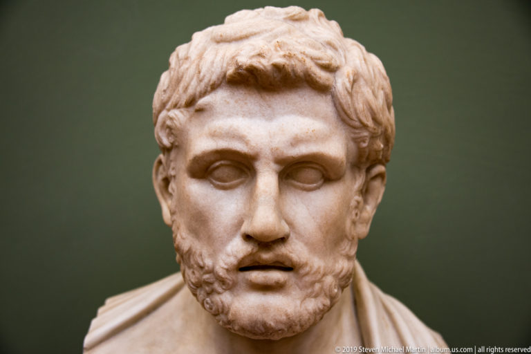 Frontal marble bust of a young bearded man with short curled hair, blank eyes, and slightly parted lips.