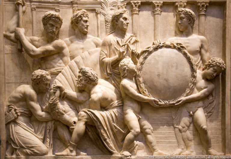 Marble relief with numerous nude and draped figures gathered around a large wreath-framed circular shield.