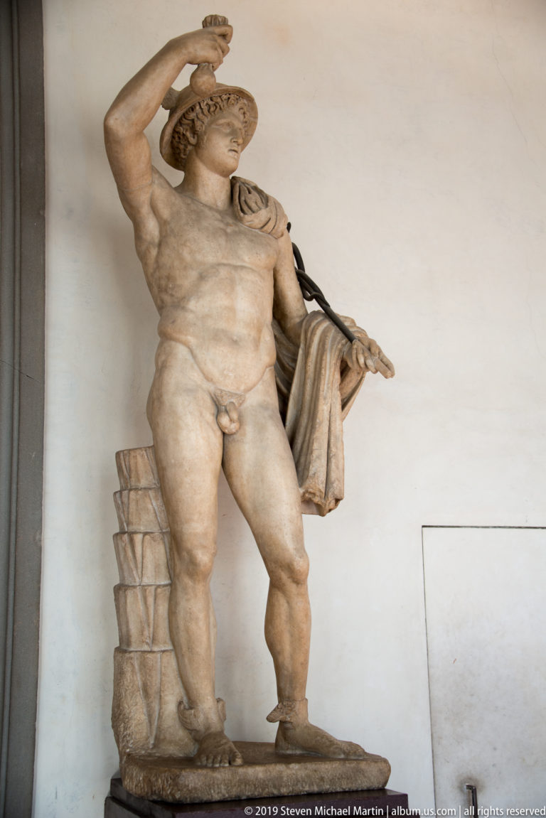 Muscular marble nude twists in a wide stance with one arm raised and drapery falling from the other.
