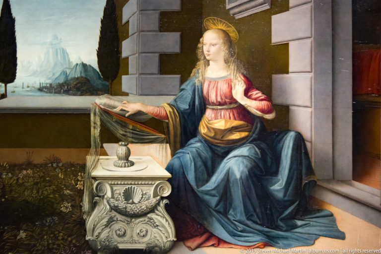 Haloed young female saint in blue and red seated beside a carved stone table, with a mountain landscape beyond.