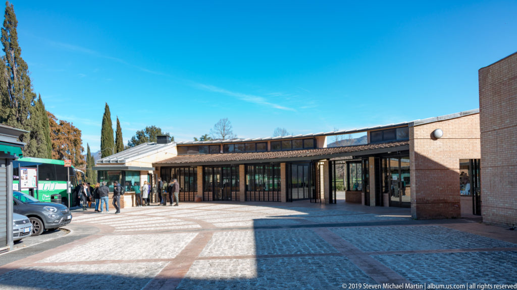 Low brick visitor entrance with glass doors and covered walkways around a paved forecourt, with visitors and vehicles nearby.