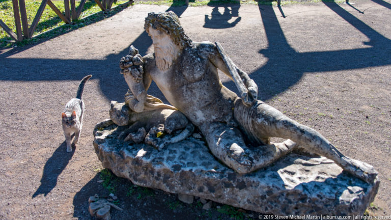 Full reclining bearded stone figure on a rough base, with a small cat walking beside the sculpture and visitor shadows behind.