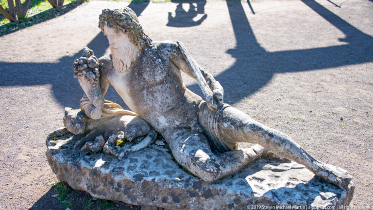 Weathered reclining bearded stone figure viewed from above on a rough base, with a clustered object raised near one shoulder.