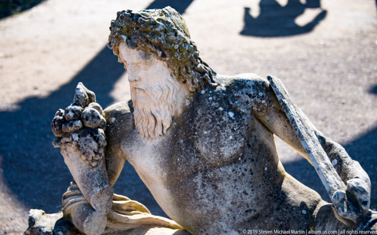 Weathered reclining bearded male stone figure holding a cluster of rounded forms near his shoulder, seen in close view.
