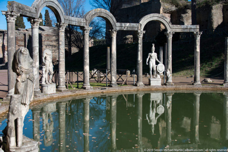 Oblique view of marble statues among columns and twin arches beside the Canopus, with broken reflections in green water.
