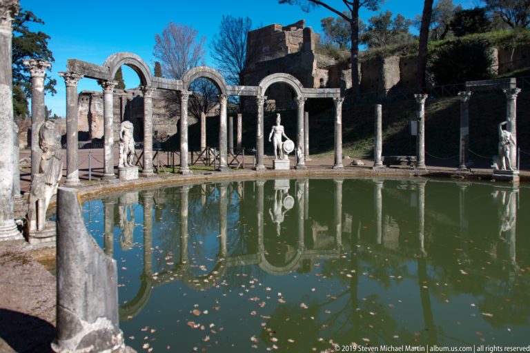 Curved green Canopus pool bordered by columns, arches, and marble statues, with sunlit brick ruins rising behind.
