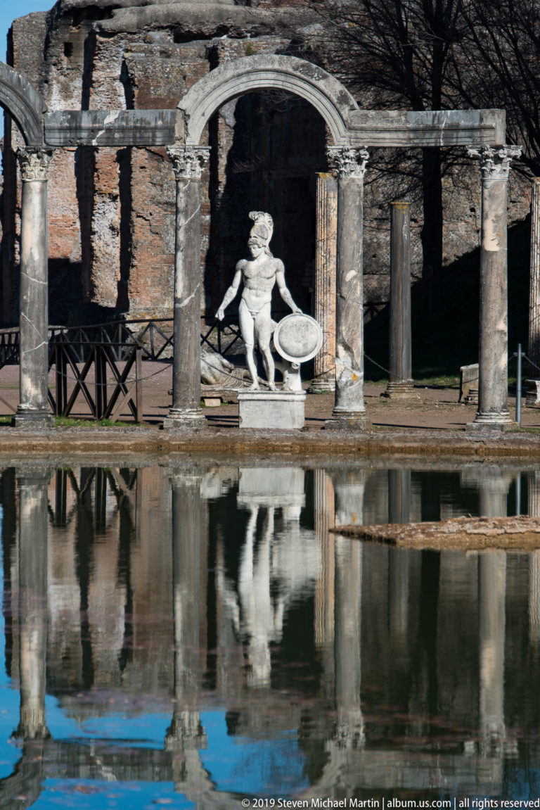 Pale reclining marble figure centered beneath a stone arch and columns beside the Canopus, reflected in the pool below.