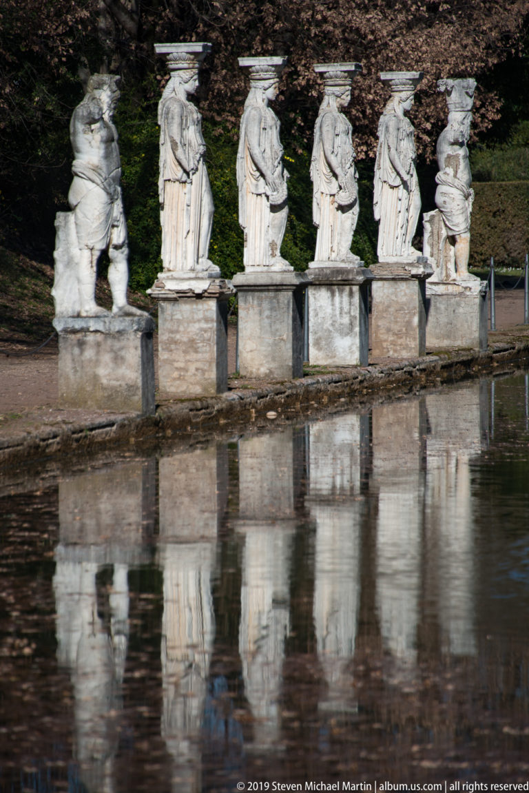 Five draped caryatid figures on square pedestals beside the Canopus, with partial reflections and a damaged figure at right.