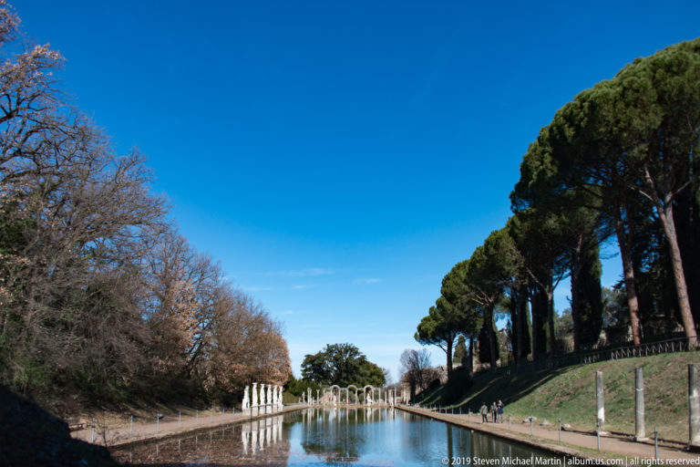 Distant view down the Canopus pool between wooded banks and umbrella pines toward white statues and an arched colonnade.