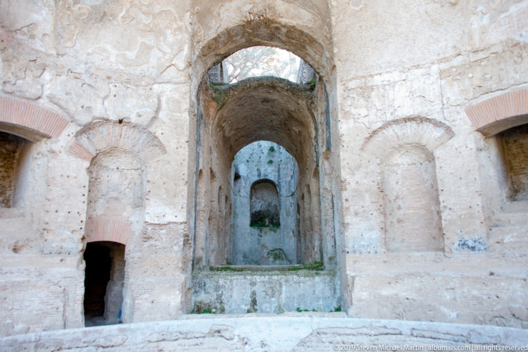 Tall vaulted opening centered in a pale Roman masonry chamber, flanked by smaller arched recesses and exposed brickwork.