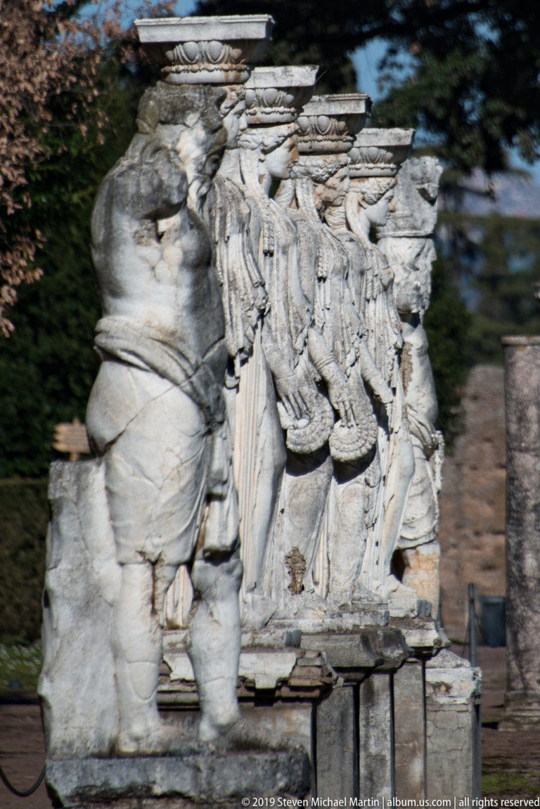 Weathered stone sculptural group with several overlapping draped figures, missing heads, and a projecting figure at left.