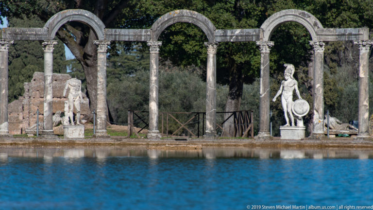Two stone arches and slender columns with shield-bearing statues along the Canopus, seen across bright blue water.