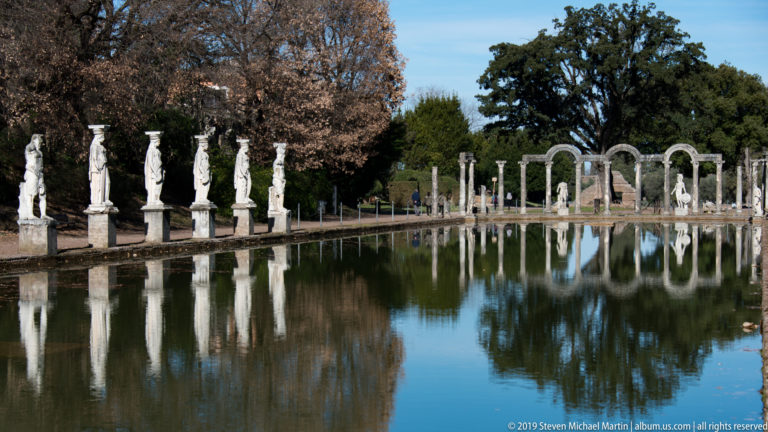 Long rectangular Canopus pool lined with white statues, with columns and arches at the far end and trees reflected in the water.