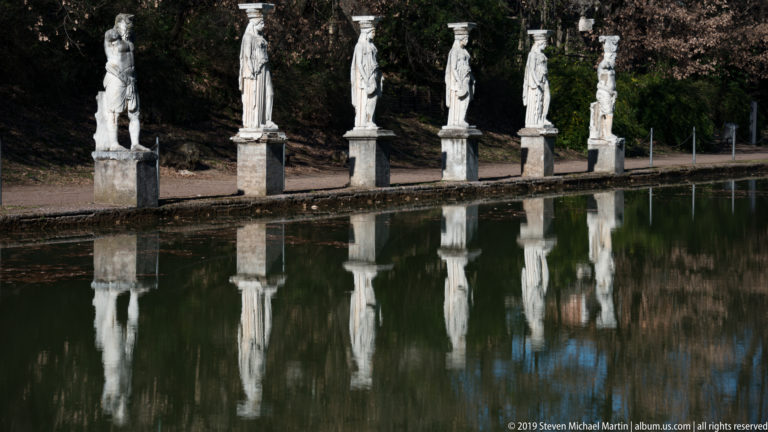 Row of five white caryatid figures on pedestals beside the Canopus, mirrored in the dark reflecting pool.