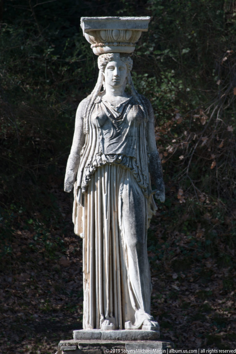 Weathered draped female caryatid figure standing frontally with an architectural block supported above her head.