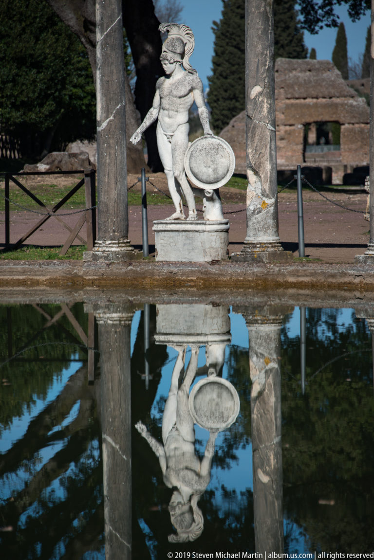 Helmeted marble figure with round shield between columns, reflected upside down in the calm Canopus pool.