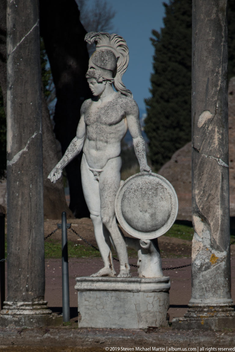 Small weathered marble figure in a crested helmet holding a round shield on a pedestal between stone columns.