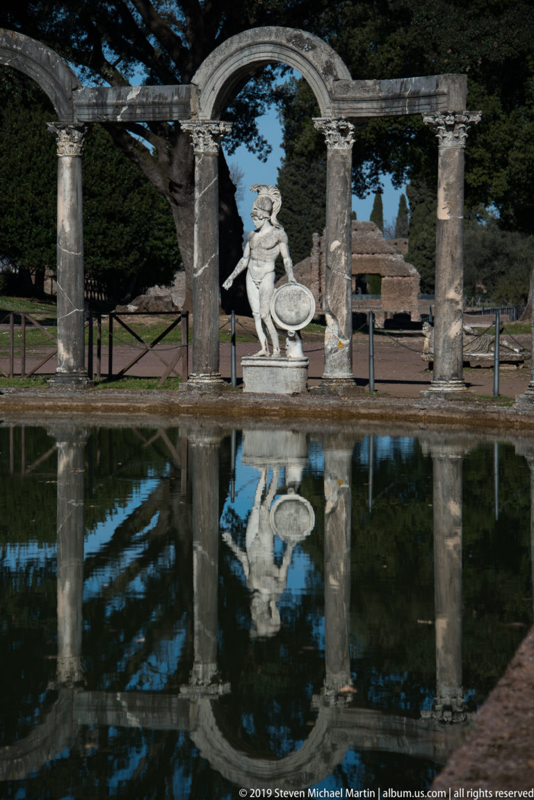 Shield-bearing marble statue centered beneath stone arches, with the figure and colonnade reflected in the Canopus pool.
