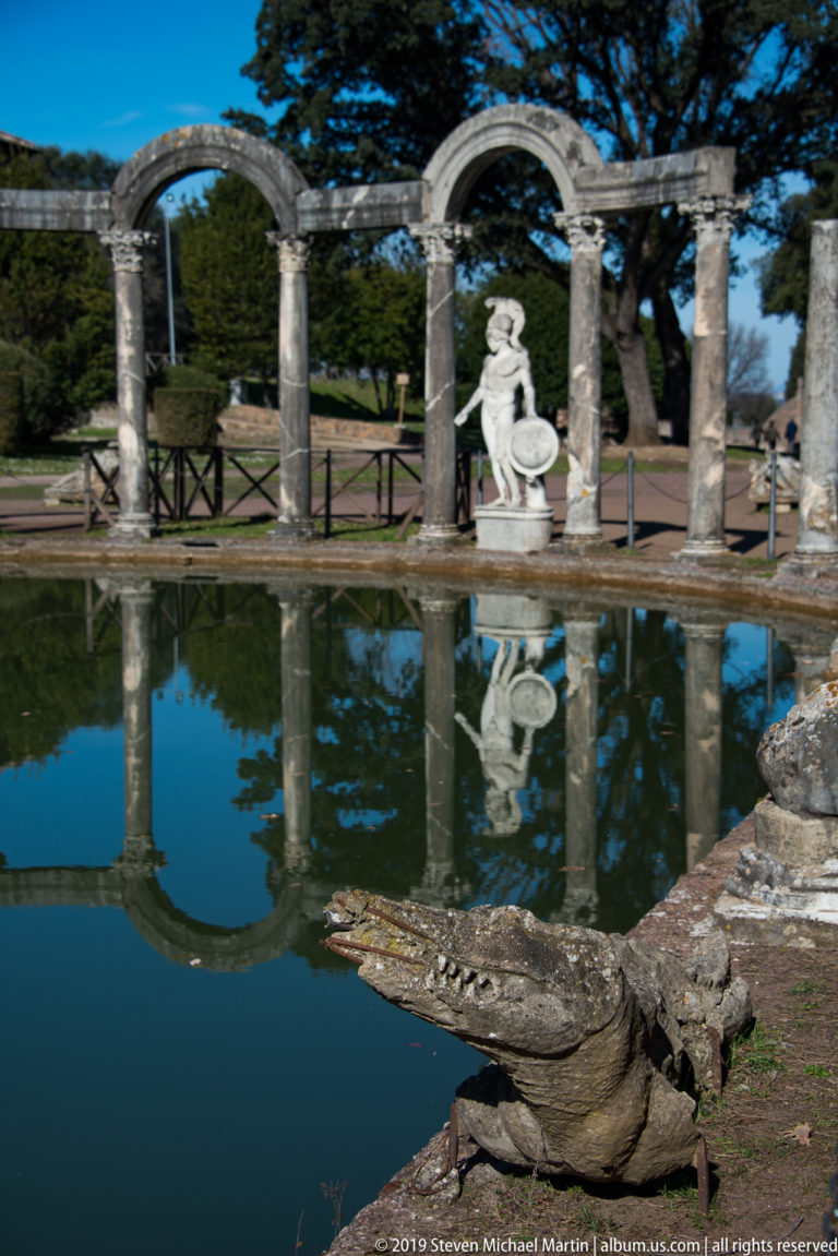 Stone crocodile sculpture beside the Canopus pool, with an arched colonnade and shield-bearing statue reflected beyond.