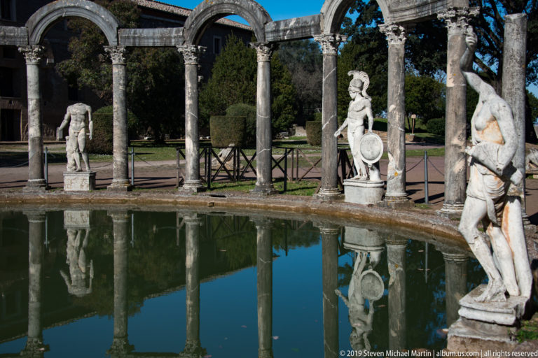 Curved stone colonnade with marble statues beside the Canopus pool, their columns and figures reflected in calm water.