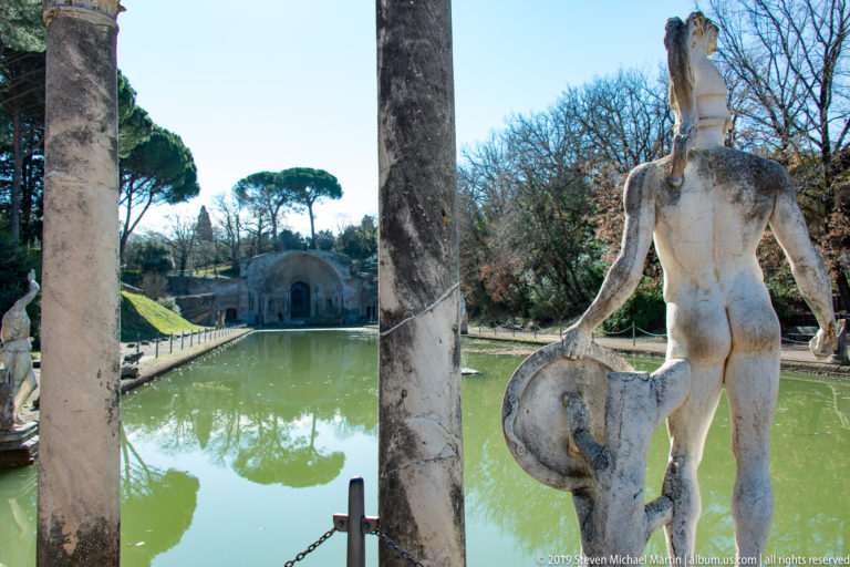 Marble shield-bearing statue between columns beside the green Canopus pool, with a vaulted structure visible at the far end.