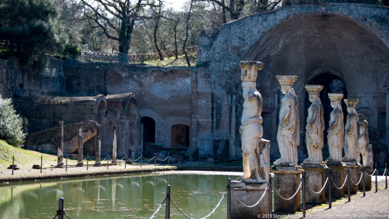 Row of pale caryatid statues beside the Canopus pool, with curved masonry and a grotto-like recess behind.