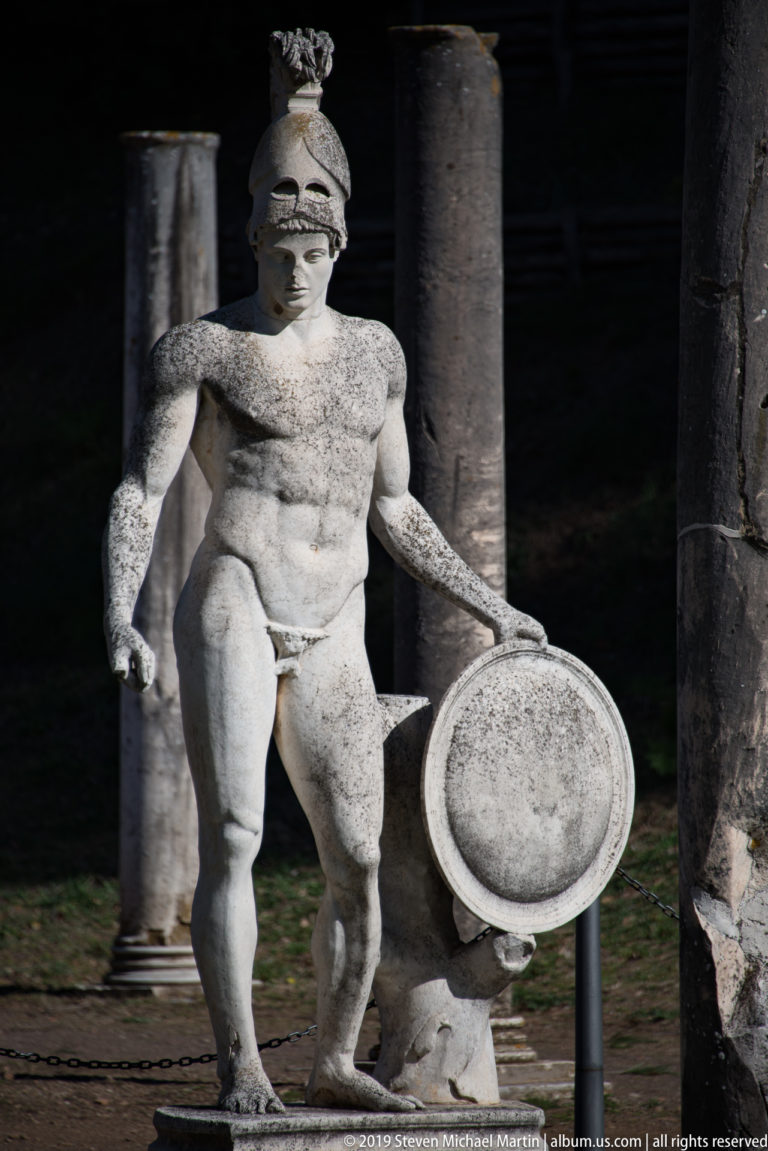 Weathered marble warrior wearing a crested helmet and holding a round shield, posed before dark stone columns.